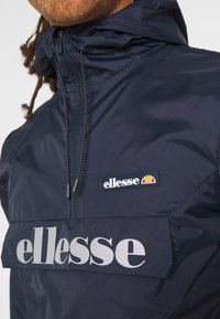 Marineblauwe windbreaker met een halve rits, voorzien van een opvallend zilveren logo en een geborduurd merkembleem, gemaakt van glad, lichtgewicht materiaal.