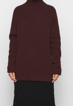 Personne portant un pull à col roulé marron foncé à manches longues et une jupe noire sur un fond clair uni.