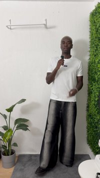 Camiseta branca com logótipo azul, calças largas pretas e sapatos pretos. Ambiente interior com planta verde e fundo de parede texturizada.