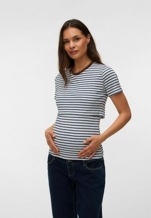 Gravid kvinde iført en stribet kortærmet bluse og mørke jeans, holder sin mave med begge hænder og står mod en ensfarvet baggrund.