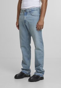 Férfi világoskék farmernadrágot visel, amelynek szegélye kopott, fekete vastag cipőkkel és fehér ujjatlan inggel, egy egyszerű szürke padlón áll.