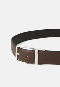 Ceinture en cuir brun avec une surface lisse, dotée d'une boucle en métal argenté et de trous d'ajustement sur toute sa longueur.