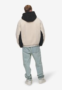 Giacca foderata in pile con corpo bianco sporco, maniche nere e cappuccio; indossata con pantaloni in denim blu chiaro e sneaker bianche.