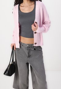 Růžový pletený cardigan s knoflíky, šedé tílko a šedé džíny s širokými nohavicemi, doplněné černou kabelkou. Viditelné texturované detaily látky.