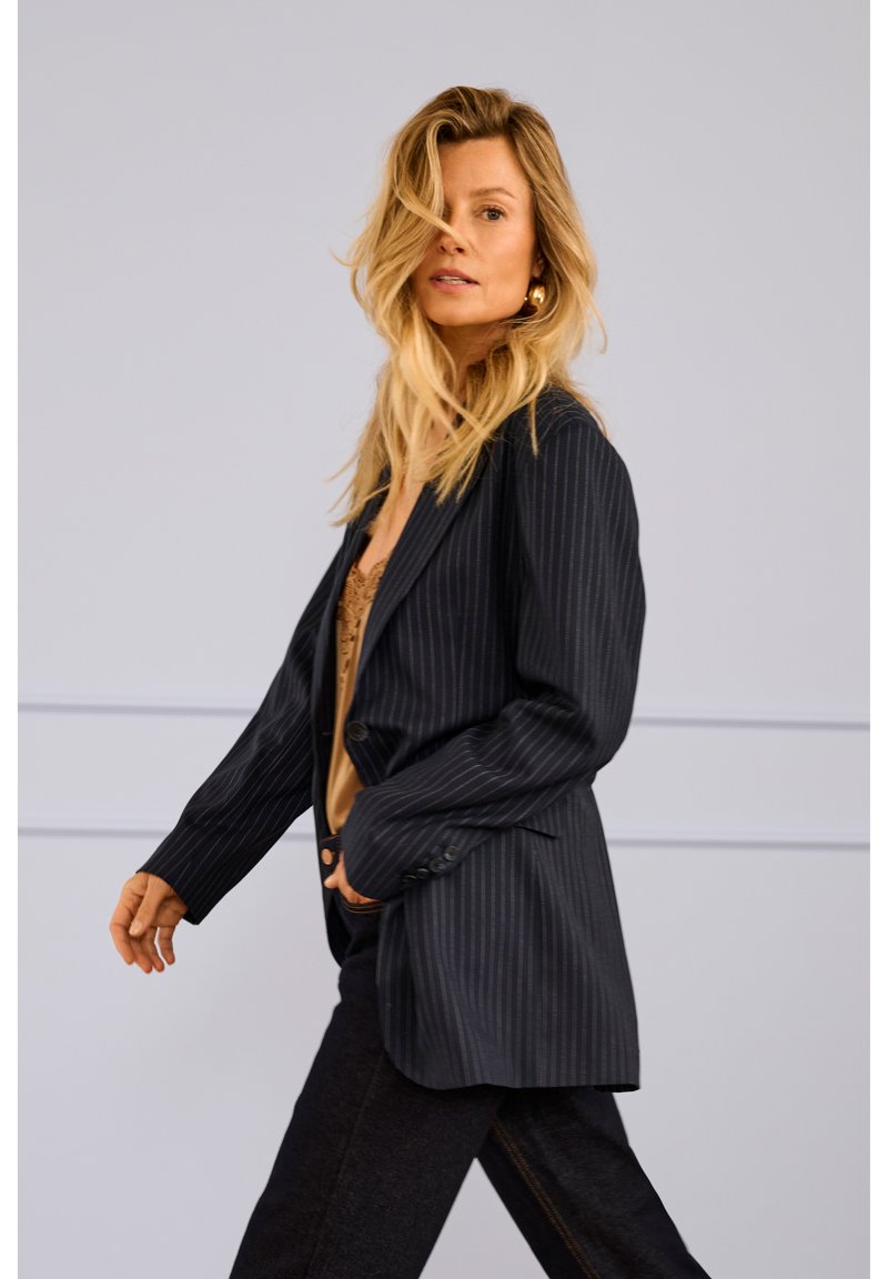 Zwarte blazer met krijtstreep, nauwsluitende pasvorm, enkelvoudige knoopsluiting en gestructureerde stof. Draagt over een beige top, gecombineerd met donkere spijkerbroek.