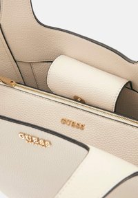 Lichtbeige handtas van gestructureerd leer, met gouden logo-accenten, een ritssluiting en contrasterende zwarte afwerking langs de randen.