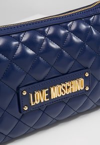 Námorníkově modrá prošívaná kožená taška se zlatou destičkou s embosovaným nápisem "LOVE MOSCHINO" a zlatými kovovými akcenty; obsahuje zipové uzavření.