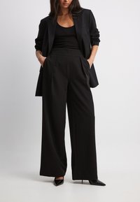 Zwarte op maat gemaakte blazer gecombineerd met wijde zwarte broek, voorzien van plooien en zijzakken. Model in zwarte puntige hakken.