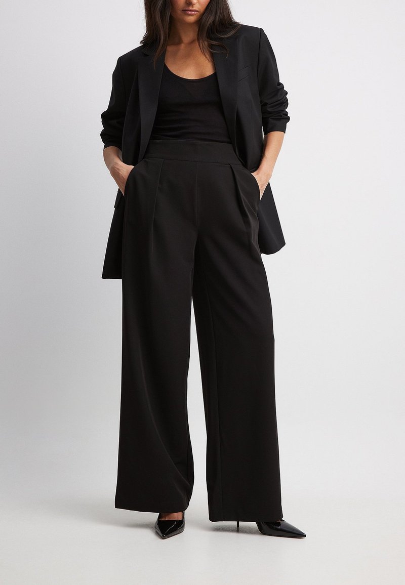 Zwarte op maat gemaakte blazer gecombineerd met wijde zwarte broek, voorzien van plooien en zijzakken. Model in zwarte puntige hakken.