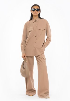 Chemise beige à boutons avec deux poches poitrine, associée à un pantalon large. Le modèle tient un sac à main rond beige et porte des lunettes de soleil noires.