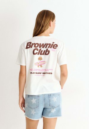 Personne portant un t-shirt blanc avec le texte "Brownie Club" et un graphisme d’un chien surfeur, associé à un short en denim bleu clair à motif d'étoiles.