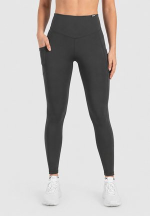 Femme portant des leggings noirs taille haute avec poches latérales et des baskets blanches sur un fond gris clair.