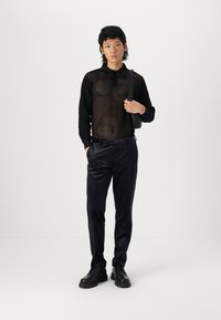 Crni mrežasti top dugih rukava, crne hlače s blagim sjajem, crne cipele i crna torbica preko tijela. Model stoji s neutralnim izrazom lica.