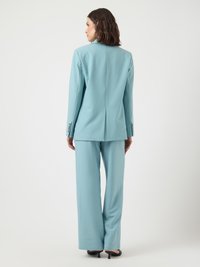 Woman in light blue tailored suit with wide-leg trousers and black heeled shoes, standing facing away on a plain white background.