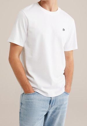 Homme portant un t-shirt blanc uni à manches courtes avec un petit logo noir brodé sur la poitrine et un jean en denim bleu clair, les mains dans les poches.