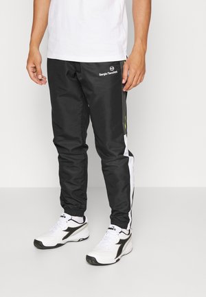 Uomo che indossa pantaloni sportivi neri Sergio Tacchini con strisce laterali bianche e grigie e scarpe sportive bianche, su sfondo neutro.