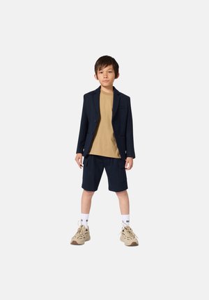 Blazer bleu marine porté sur un t-shirt beige, associé à un short bleu marine et des baskets beiges. Comprend des chaussettes sportives et une coupe décontractée.