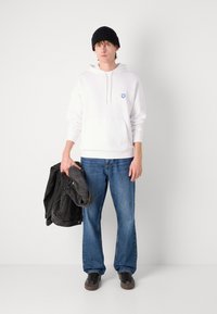Felpa bianca con tasca frontale e emblema blu, abbinata a jeans in denim blu e una giacca di denim nera tenuta in mano, completata da un berretto nero.