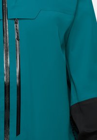 Türkisjacke mit schwarzem, wasserfestem Frontreißverschluss und vertikalem, gesichertem Brusttaschenreißverschluss, mit schwarzem Bündchendetail an den Ärmeln.