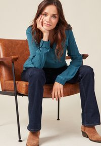 Camicia button-up turchese, jeans a zampa d'elefante in denim scuro e stivaletti color cammello. Seduto su una sedia in pelle marrone con braccioli in metallo.