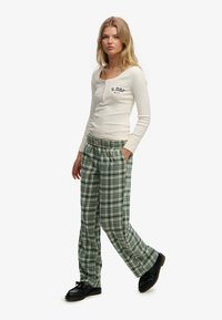 Long-sleeve cream shirt with button placket and logo; green plaid pants with wide legs and elastic waistband; paired with black shoes.