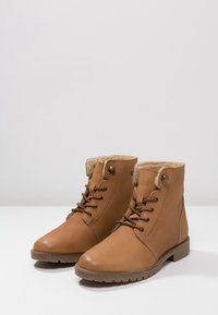 Bottines en cuir marron avec doublure en peluche, devant à lacets, bout rond et semelle en caoutchouc texturée. Comprend des œillets en métal et un détail en bouton.
