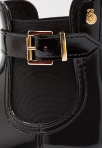 Bottines noires en cuir verni avec panneau élastique, détail de boucle dorée et emblème doré. Texture lisse et brillante avec des bords cousus.