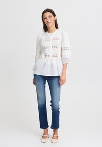 Camicia bianca a maniche lunghe con dettagli in pizzo, orlo peplum e chiusura a bottoni, abbinata a jeans blu a gamba dritta e scarpe basse beige.