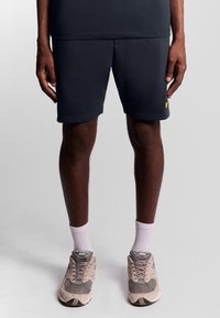 Donker marineblauwe shorts met een gladde textuur, voorzien van een gele logo-accent. Gecombineerd met beige sneakers en witte enkelsokken.