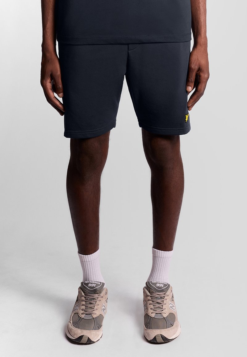 Donker marineblauwe shorts met een gladde textuur, voorzien van een gele logo-accent. Gecombineerd met beige sneakers en witte enkelsokken.
