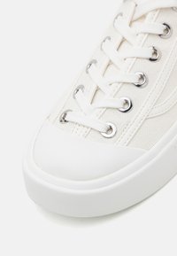 Gros plan sur la pointe d'une basket en toile blanche avec des lacets blancs passés à travers des œillets métalliques et un bord de semelle en caoutchouc.