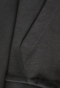 Tissu gris foncé, tricot texturé avec une finition lisse. Présente des coutures visibles et un tombé naturel, avec des sections de matériau qui se chevauchent.
