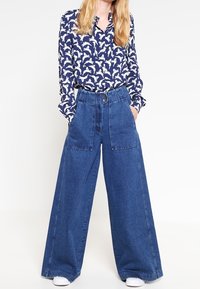 Camicetta navy con motivo di uccelli bianchi, colletto abbottonato, abbinata a pantaloni in denim blu a gamba larga con grandi tasche e vita alta.