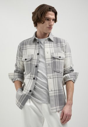Camicia a quadri grigia e bianca con due tasche sul petto, realizzata in un tessuto testurizzato, indossata sopra una t-shirt grigia chiara.