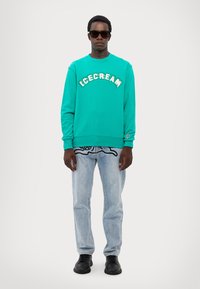 Felpa turchese con stampa "ICECREAM", abbinata a jeans denim blu chiaro con un design a contorno nero e scarpe nere.