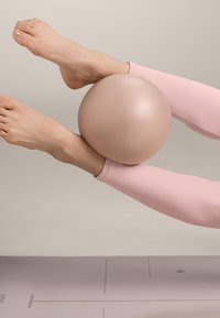 Picioare cu colanți roz țin o minge de exerciții bej între picioare deasupra unei saltele de yoga texturate pe un fundal neutru.