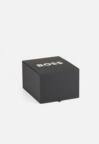 Caja negra cuadrada hecha de cartón, con un logo blanco de "BOSS" en la parte superior. Color sólido con una textura suave. Base rectangular, parte superior abierta.