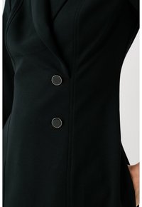 Robe noire ajustée avec un décolleté drapé, une texture lisse, et deux boutons noirs proéminents avec des accents dorés sur le côté.