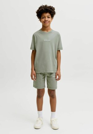 Chlapec s kučeravými vlasmi, oblečený v tričku a kraťasoch v zelenej šalviovej farbe, bielych ponožkách a bielych teniskách, stojí na jednofarebnom pozadí.