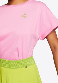 Person med en rosa kortärmad skjorta med liten grön palmträdbroderi och en ljusgrön kjol med en texturerad midja.