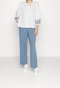 Blouse en coton blanc avec des accentuations noires brodées sur les manches bouffantes, associée à un pantalon large bleu clair et des baskets blanches.