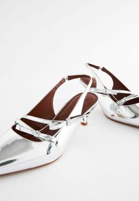 Paire de chaussures slingback argentées brillantes à talons bas, bouts pointus, fines brides et petits nœuds décoratifs sur fond blanc.