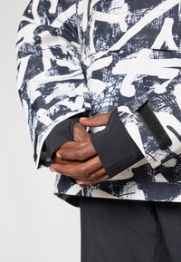 Μαύρο και λευκό σαρίκι με μοτίβο, μακριά μανίκια, ρυθμιζόμενους καρπούς και λείο υλικό. Χέρια τοποθετημένα μαζί μπροστά.