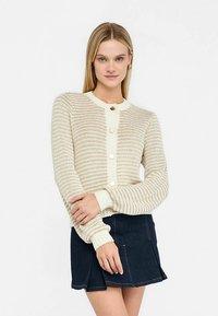 Gestrickte Strickjacke in Creme mit strukturierten Streifen, mit rundem Ausschnitt, Knopfverschluss und gerippten Bündchen. Kombiniert mit einem dunkelblauen Jeansrock.