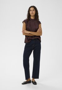 Tmavohnedý top s krémovými bodkami a krátkymi rukávmi, v kombinácii s vysokým pásom tmavomodrých džínsov a čiernymi roztiahnutými topánkami.