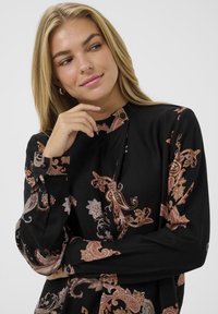 Zwarte blouse met een paisleypatroon in tinten van koper en zilver. Lange mouwen, hoge halslijn en knoopaccenten op de kraag. Gladde stof.