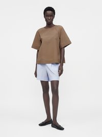 Femme debout portant un haut marron à manches courtes, un short bleu clair et des ballerines noires sur un fond blanc uni.