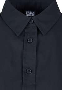 Camicia nera con colletto a medium spread, cinque bottoni neri in plastica e una trama liscia; etichetta visibile all'interno del colletto.