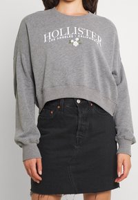 Sweat-shirt gris raccourci avec le texte blanc "Hollister" et des accents floraux, associé à une jupe en denim noir ajustée avec un ourlet effiloché.
