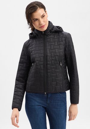 Frau trägt eine schwarze gesteppte Reißverschlussjacke mit Kapuze und blaue Jeans, steht vor einem einfarbigen hellen Hintergrund.